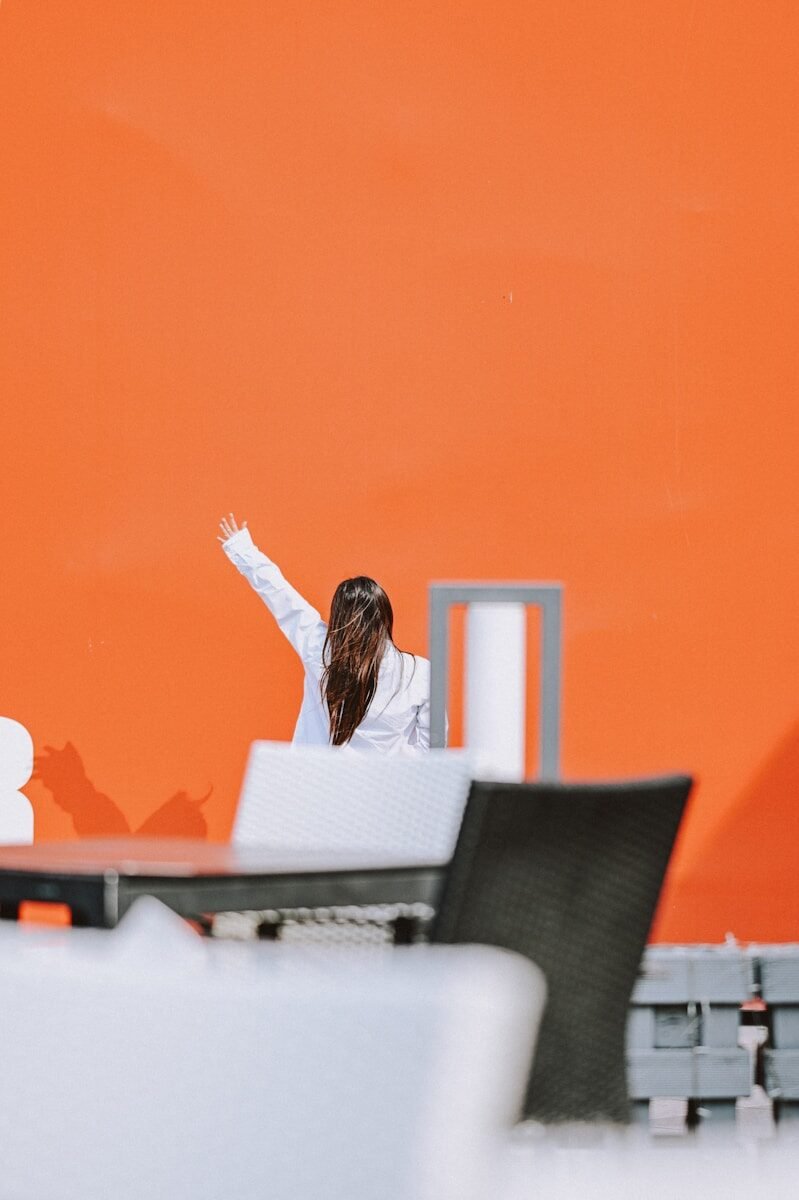 Woman in white jacket with arm raised against orange wall